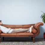 Adult man wearing a blue shirt and beige pants lying down on psychiatrist couch. The background is blue wall. His eyes are closed. Shot indoor with a full frame DSLR camera.